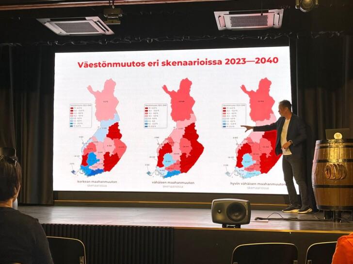 Tulevaisuuspäivästä otettu kuva, jossa puhuja osoittaa takana olevaa esitystä. Esityksessä on näkyvillä väestönkehitys ennuste.