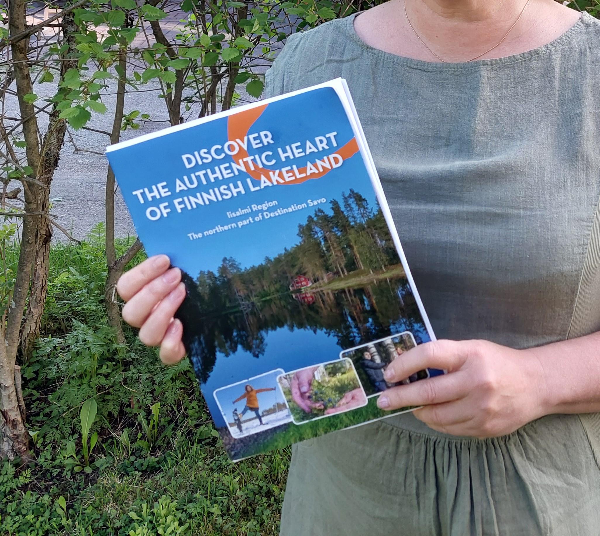 Esitettä pitää kädessään nainen. Esitteen kansikuvassa on suomalainen järvimaisema kesällä.